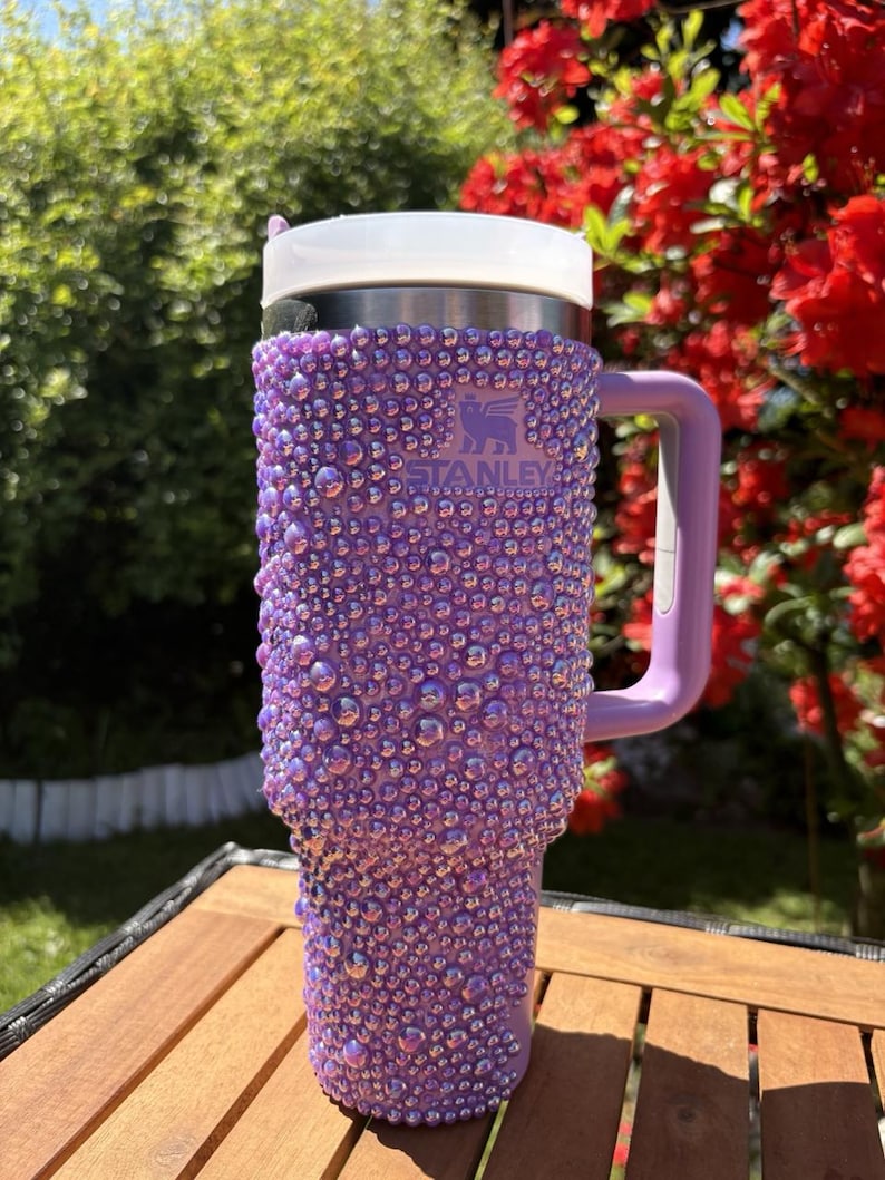 May include: A purple Stanley tumbler covered in iridescent rhinestones. The tumbler has a white lid and a purple handle. The Stanley logo is visible on the front. The tumbler is sitting on a wooden table outdoors.