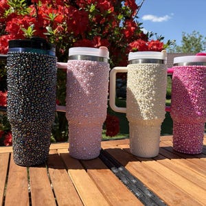 May include: Four insulated tumblers with handles, each covered in decorative beads. Colors include black, light pink, cream, and hot pink. The tumblers are arranged on a wooden surface with a backdrop of red flowers and a blue sky.