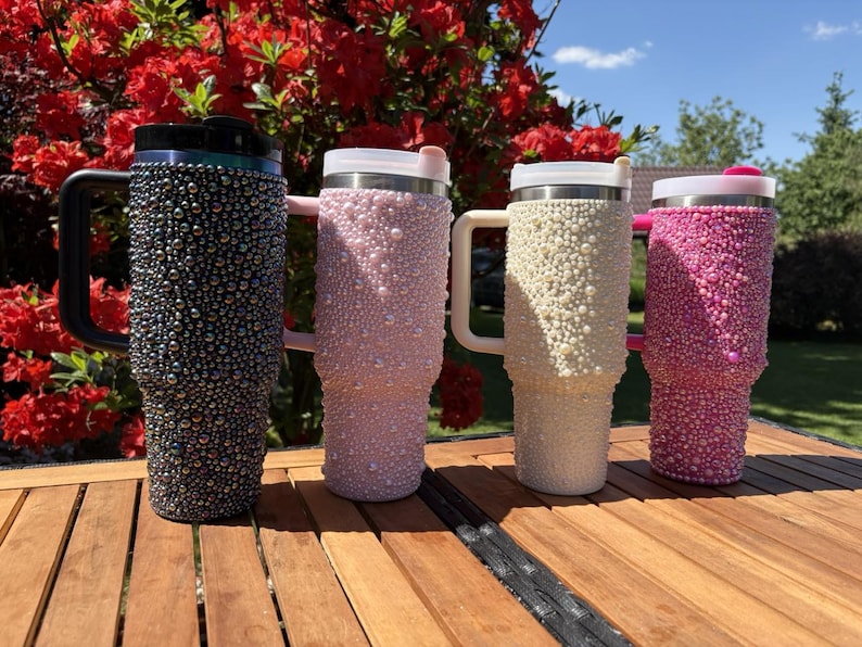 May include: Four insulated tumblers with handles, each covered in decorative beads. Colors include black, light pink, cream, and hot pink. The tumblers are arranged on a wooden surface with a backdrop of red flowers and a blue sky.