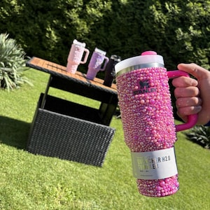 May include: A pink Stanley Quencher H2.0 Tumbler covered in small, sparkling embellishments. The tumbler has a pink handle and a white and silver lid. Other tumblers in pink, purple, and black are on a table in the background.