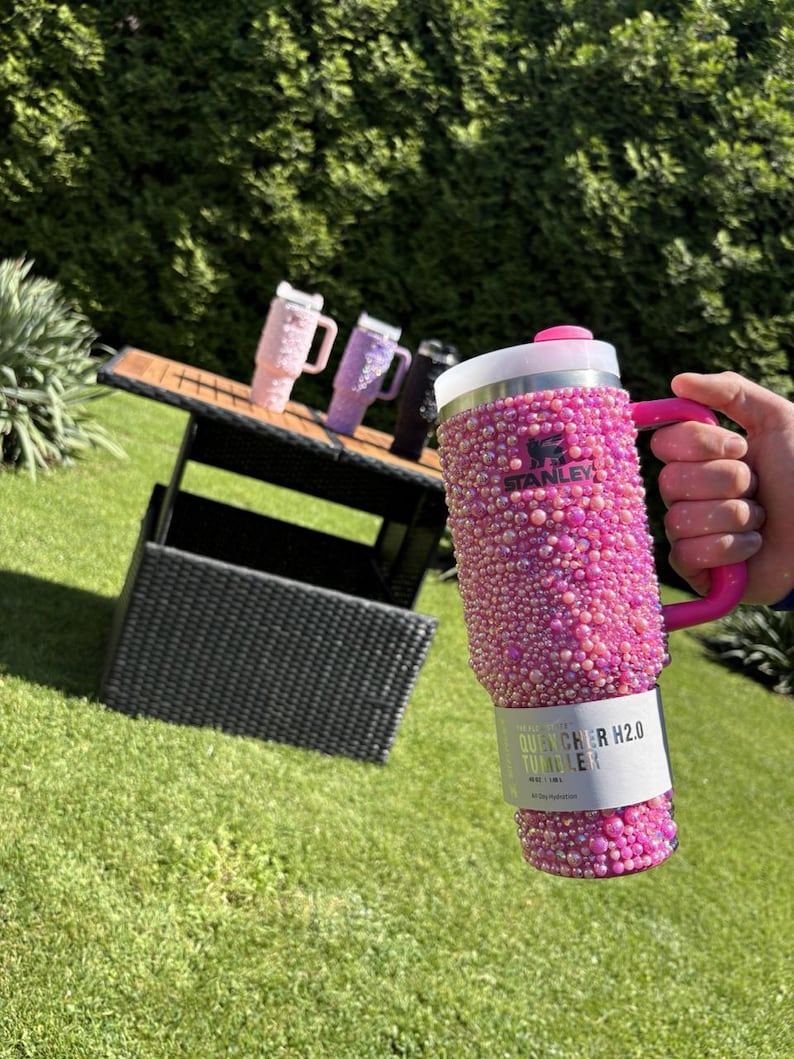 May include: A pink Stanley Quencher H2.0 Tumbler covered in small, sparkling embellishments. The tumbler has a pink handle and a white and silver lid. Other tumblers in pink, purple, and black are on a table in the background.