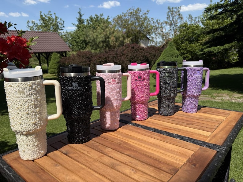 May include: Six bedazzled Stanley tumblers in a row, each with a different color scheme. Colors include white, black, pink, dark pink, grey, and purple. Each tumbler has a handle and a lid. The tumblers are sitting on a wooden table.