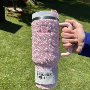 May include: A pink Stanley Quencher H2.0 tumbler, embellished with numerous pink pearls. The tumbler has a handle and a white lid. The Stanley logo is visible. The tumbler is held against a green background.