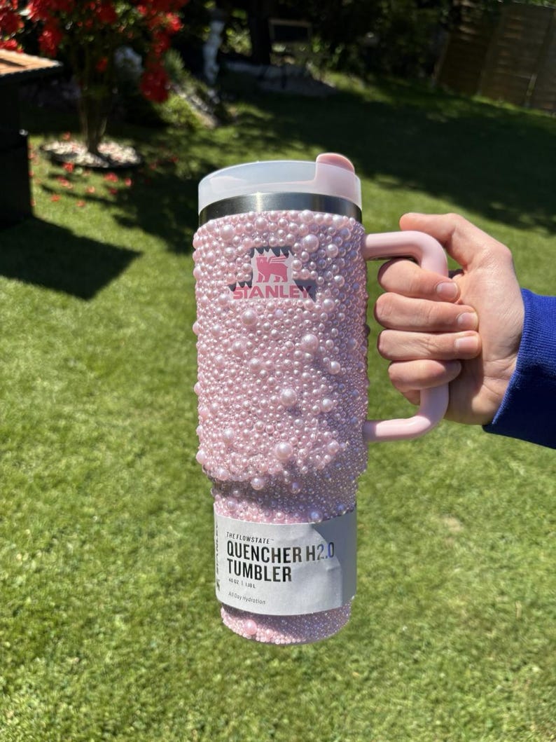 May include: A pink Stanley Quencher H2.0 tumbler, embellished with numerous pink pearls. The tumbler has a handle and a white lid. The Stanley logo is visible. The tumbler is held against a green background.