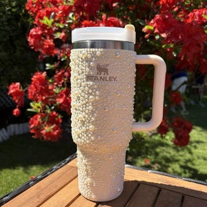 May include: A cream-colored Stanley tumbler, embellished with faux pearls and rhinestones. The tumbler has a white handle and lid, with the Stanley logo visible. The background features red flowers and a wooden surface.