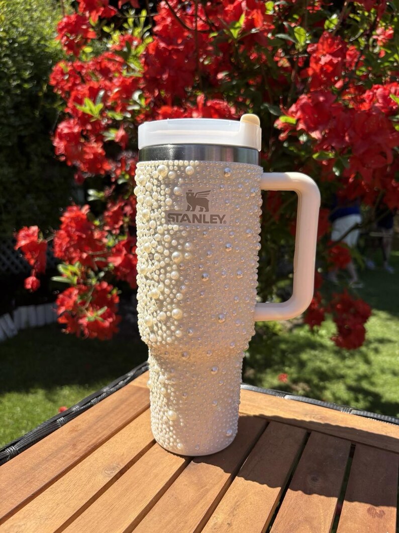 May include: A cream-colored Stanley tumbler, embellished with faux pearls and rhinestones. The tumbler has a white handle and lid, with the Stanley logo visible. The background features red flowers and a wooden surface.