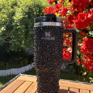 May include: Black Stanley travel mug with a textured, bubble-like surface. The mug has a black handle and lid, and the Stanley logo is visible. The background features green foliage and red flowers, suggesting an outdoor setting.