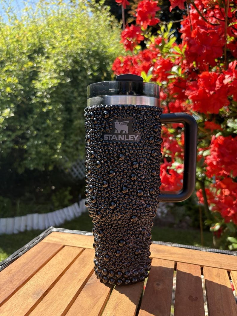 May include: Black Stanley travel mug with a textured, bubble-like surface. The mug has a black handle and lid, and the Stanley logo is visible. The background features green foliage and red flowers, suggesting an outdoor setting.