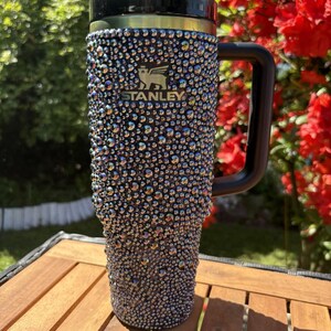 May include: A bedazzled Stanley travel mug with a black handle and lid. The mug is covered in iridescent rhinestones, creating a sparkling effect. The Stanley logo is visible on the front. The mug sits on a wooden table, with greenery and red flowers in the background.