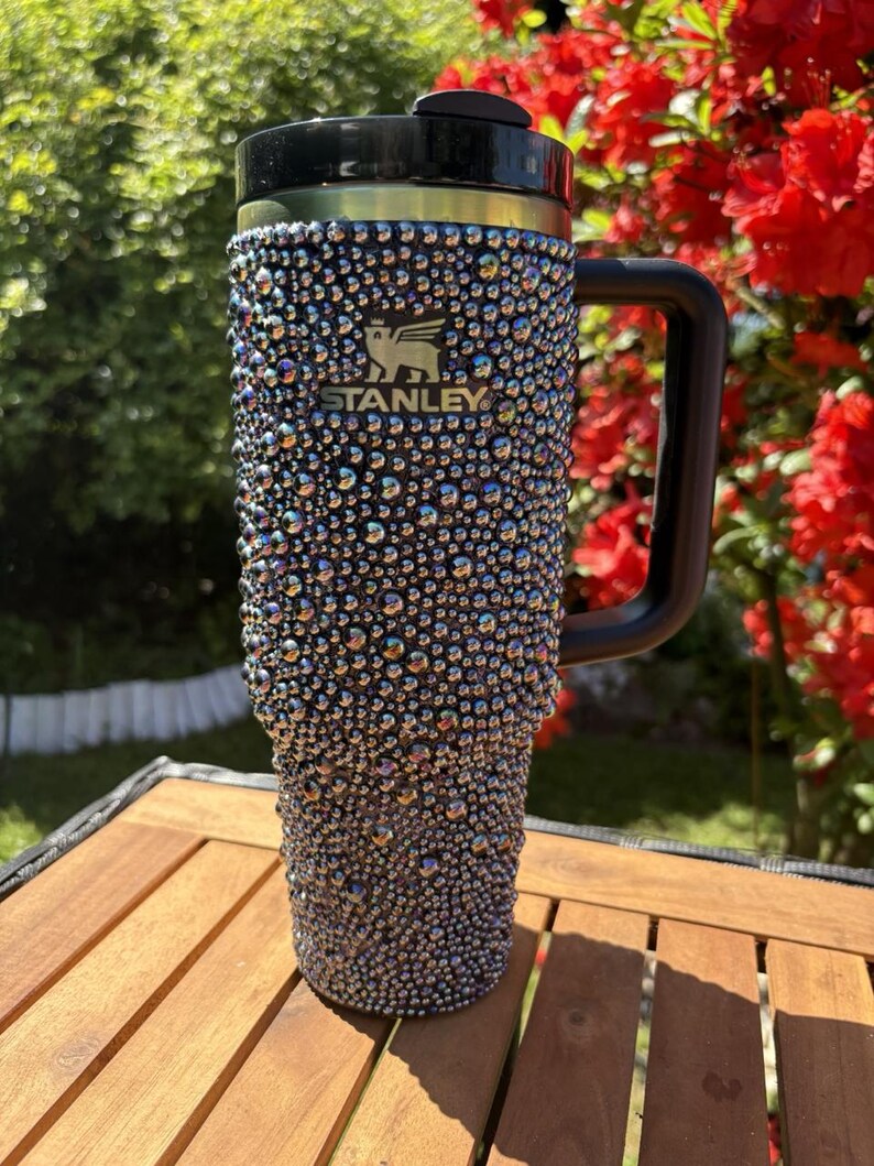 May include: A bedazzled Stanley travel mug with a black handle and lid. The mug is covered in iridescent rhinestones, creating a sparkling effect. The Stanley logo is visible on the front. The mug sits on a wooden table, with greenery and red flowers in the background.