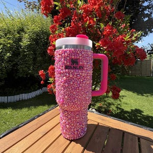 May include: A pink Stanley tumbler covered in iridescent rhinestones. The tumbler has a pink handle and a white and pink lid. The Stanley logo is visible on the front. The tumbler is sitting on a wooden table.