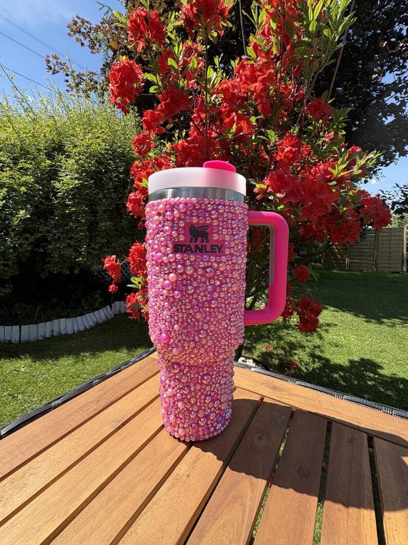 May include: A pink Stanley tumbler covered in iridescent rhinestones. The tumbler has a pink handle and a white and pink lid. The Stanley logo is visible on the front. The tumbler is sitting on a wooden table.