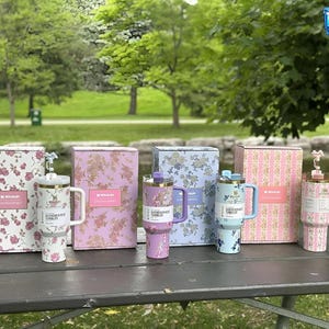 May include: A white ceramic mug with a black handle and the text "But First, Coffee" in a black, handwritten-style font. The mug is on a white surface.