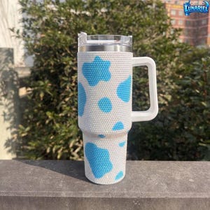 May include: A white and blue tumbler with a handle. The tumbler is covered in white rhinestones with blue cow spots. The tumbler has a clear lid and a white handle. The brand name "LUNARIEL" is visible in the top right corner.