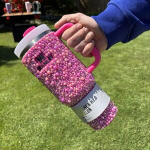 May include: A pink Stanley H2.0 Flowstate tumbler covered in pink rhinestones. The tumbler has a pink handle and a white lid. The text "Stanley" is visible on the side. The tumbler is held by a person.