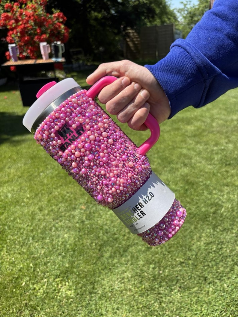 May include: A pink Stanley H2.0 Flowstate tumbler covered in pink rhinestones. The tumbler has a pink handle and a white lid. The text "Stanley" is visible on the side. The tumbler is held by a person.