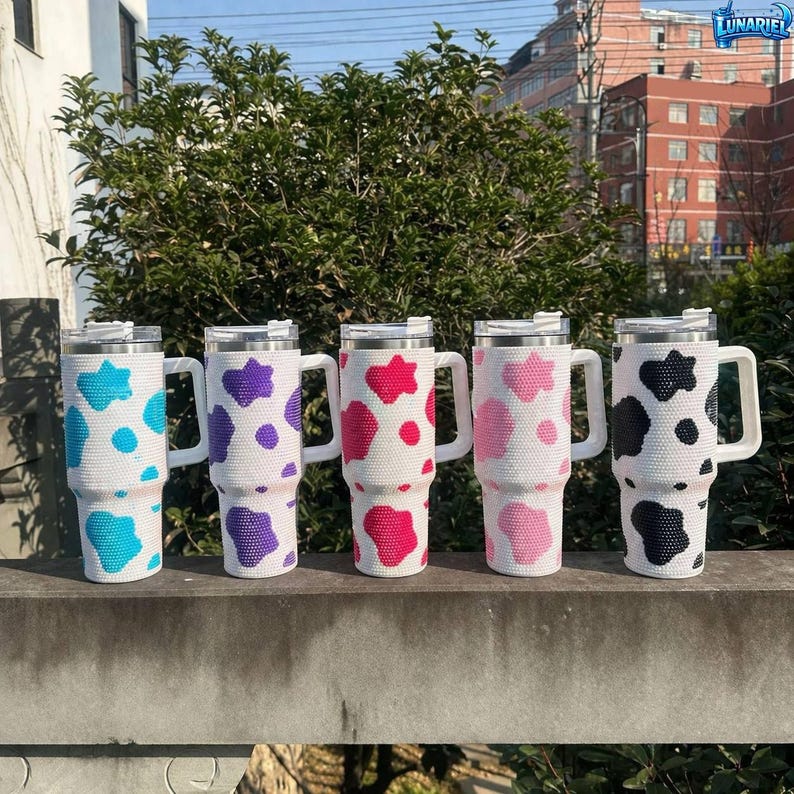 May include: Five white insulated travel mugs with handles, each adorned with a different color cow print pattern: turquoise, purple, red, pink, and black. The mugs have silver lids and white handles.