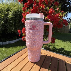 May include: A pink Stanley tumbler, embellished with numerous pink pearls, features a white lid and a matching pink handle. The tumbler is set on a wooden table, with a backdrop of vibrant red flowers and green foliage.