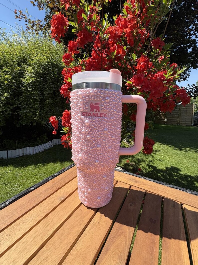 May include: A pink Stanley tumbler, embellished with numerous pink pearls, features a white lid and a matching pink handle. The tumbler is set on a wooden table, with a backdrop of vibrant red flowers and green foliage.