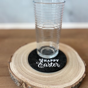 Könnte beinhalten: Ein klares Glas steht auf einem schwarzen Filzuntersetzer mit dem weißen Schriftzug "Happy Easter". Der Untersetzer liegt auf einer natürlichen Holzscheibe mit sichtbarer Maserung.