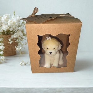 May include: A brown cardboard gift box with a clear window showing a white puppy-shaped candle. The box has a decorative leaf-shaped closure and the text "Aydas Aromatics". A small bouquet of white flowers is in the background.