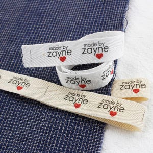 Puede incluir: Primer plano de varias etiquetas de tela blancas y beige con el texto "made by zayne" en negro, cada una con un pequeño corazón rojo. Las etiquetas están dispuestas sobre una tela azul y blanca.