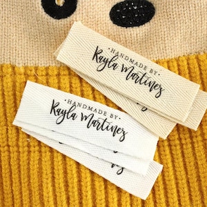 以下が含まれることがあります： 「HANDMADE BY Kayla Martinez」の黒い筆記体で書かれた2つの布製ラベルのクローズアップ。ラベルは白とオフホワイトで、織り目加工が施されています。背景は黄色のリブ編み生地です。