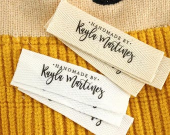 Peut inclure: Gros plan sur deux étiquettes en tissu avec le texte "HANDMADE BY Kayla Martinez" en écriture noire. Les étiquettes sont blanches et écrus, avec un tissage texturé. Le fond est un tissu côtelé jaune.