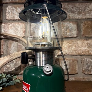 May include: A green Coleman lantern with a clear glass globe, a dark green metal top, and a silver metal handle. The base features a red and white Coleman logo, a black control knob, and a silver fuel cap. A vertical LED bulb is visible inside.