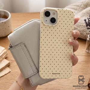 May include: A cream-coloured phone case with a black polka dot pattern is held next to a light grey wallet. The phone case has two camera lenses and the word "Tomorrow" printed at the bottom. The case is from Markcraft Cases Studio.