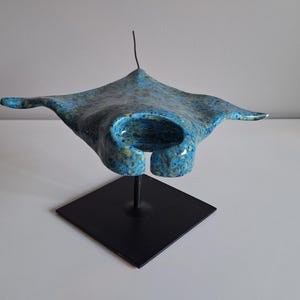 May include: A ceramic manta ray sculpture with a blue and turquoise mottled glaze. The sculpture is mounted on a black metal stand with a square base. The ray's wings are spread wide, creating a decorative piece. The sculpture is a decorative item.