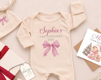 May include: A cream-colored ribbed long-sleeve bodysuit with the text Sophia's FIRST CHRISTMAS 2026 and a pink bow graphic, a matching ribbed hat with a pink bow, a white gift box with a red ribbon, a gift tag, and a Merry Christmas card.