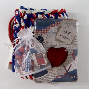 May include: A collection of hockey-themed items. Includes a phone case with a collage design, a red heart-shaped object, a small bag of candies, and a braided scarf in red, white, and blue. The phone case has the name "Graham" and the number "44".