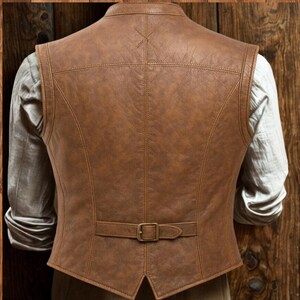 May include: A brown leather vest with a classic design. The vest features a notched collar, a buckle closure at the back, and detailed stitching. The vest is shown from the back, with a light-colored shirt visible underneath.