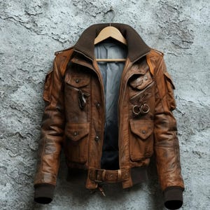 May include: A brown leather jacket with multiple pockets, a collar, and a belt. The jacket is hanging on a wooden hanger against a textured grey wall. The jacket has a bomber style with a ribbed hem and cuffs.