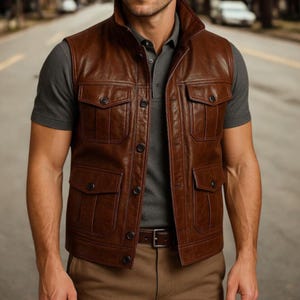 May include: A brown leather vest featuring four front pockets and a button-up design. The vest has a high collar and is worn over a grey polo shirt. A brown belt is visible at the waist. The vest appears to be made of high-quality leather.