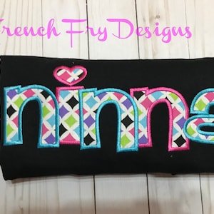 May include: Black fabric with the name "Nina" embroidered in colorful, patterned letters. The letters are outlined in blue and filled with a geometric design in pink, green, purple, and black. A pink heart with a geometric pattern sits above the "i".
