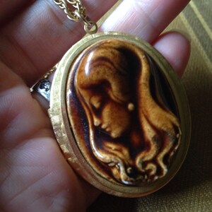 May include: A gold chain necklace with a locket pendant featuring a brown and white ceramic cameo of a woman's face.