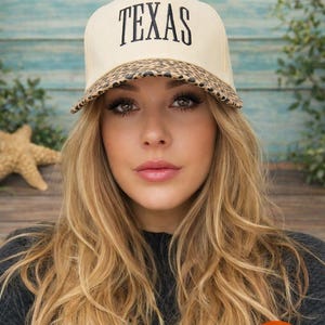May include: A beige baseball cap featuring the word TEXAS embroidered in black thread on the front panel. The brim of the cap is decorated with a brown and black leopard print pattern. The cap is structured with a curved visor and a solid crown.