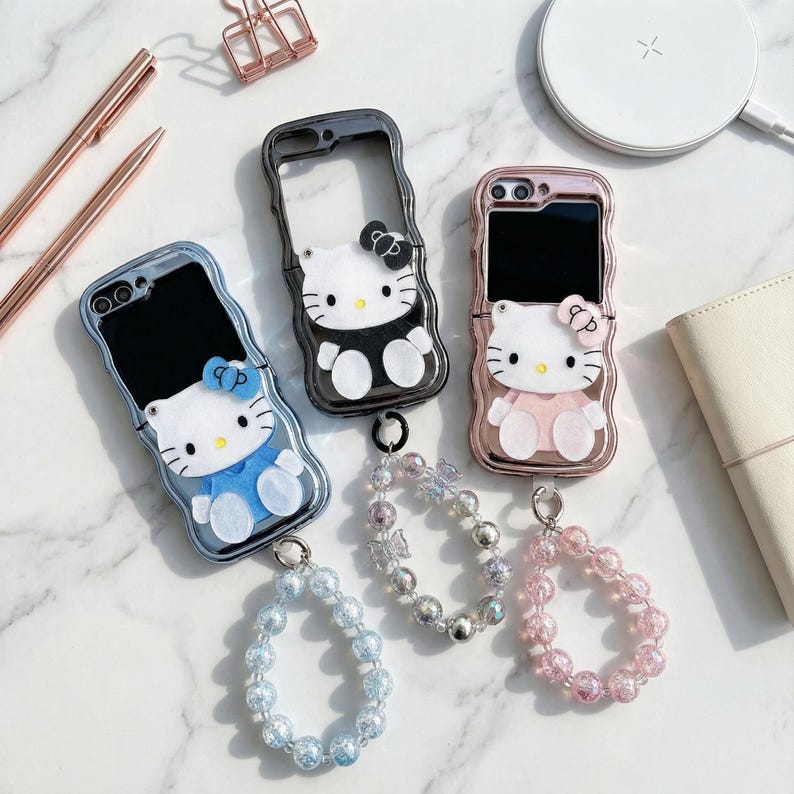 May include: A collection of phone cases featuring a Hello Kitty design. The cases are in various colors, including blue, black, and rose gold. Each case has a matching beaded wrist strap. The cases are on a white marble surface.