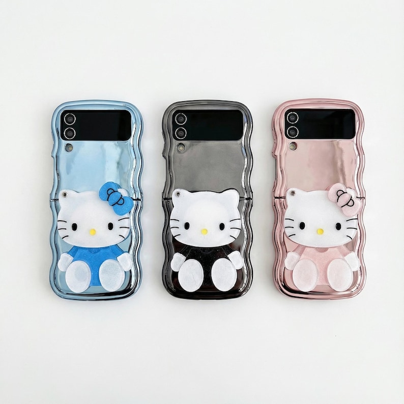 May include: Three phone cases featuring a Hello Kitty design. The cases are in blue, gray, and pink, each with a different outfit for the character. The cases have a glossy finish and are designed to fit a foldable phone.