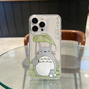 May include: Clear phone case featuring a cartoon Totoro character holding a leaf umbrella. The design includes a gray Totoro, a smaller Totoro, and green foliage. The word "TOTORO" is printed vertically on the side.