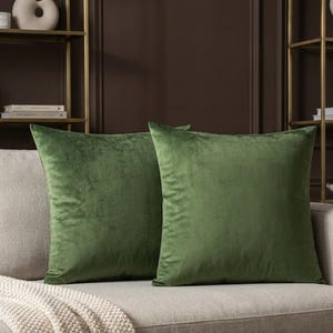 May include: Two square, olive green velvet cushions sit on a beige sofa. A cream-coloured knitted throw blanket is draped over the sofa. The cushions add a pop of colour to the neutral-toned living room.