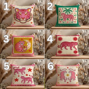May include: A collection of six decorative cushions, each with a pink tiger design. The cushions feature various tiger illustrations on cream or coloured backgrounds. Designs include tiger faces, tigers with moons, and tigers with stars. The cushions have pink borders.