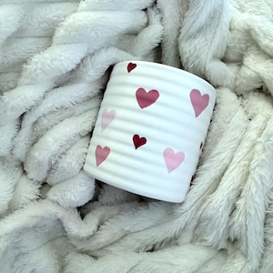 Puede incluir: Jardinera cilíndrica de cerámica blanca con textura acanalada, adornada con corazones rosas. La jardinera está sobre una manta suave y blanca. Un artículo decorativo para el hogar.