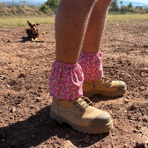 May include: A person wearing tan work boots and pink leg gaiters with a pizza slice pattern. A dog is in the background. The gaiters are gathered at the top and cover the ankles.