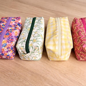 Pode incluir: Quatro estojos de tecido com diferentes padrões. O primeiro é coberto por flores roxas, azuis e amarelas com um zíper roxo. O segundo tem um fundo branco com folhas verdes e um zíper verde. O terceiro é amarelo xadrez com um zíper branco. O último é rosa com fatias de pizza e um zíper rosa.