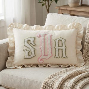 May include: Cream-colored rectangular pillow with ruffled edges. The pillow features the embroidered letters "SDA" in green and pink. The pillow is placed on a beige sofa with a knitted throw.