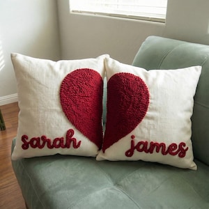 May include: Two white cushions with a red, textured heart design. One cushion has the name "sarah" embroidered in red, and the other has "james". The cushions are on a light green sofa.