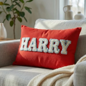 Puede incluir: Un cojín rectangular rojo con el nombre "HARRY" bordado en blanco, delineado con azul claro. El cojín está colocado en un sofá gris, con una manta color crema sobre el brazo.